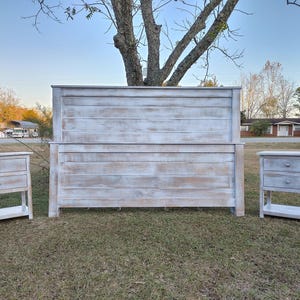 May include: A distressed white wooden bedroom set, including a headboard and two nightstands. The headboard is the focal point, flanked by the matching nightstands with two drawers each. The set is placed on a grassy lawn.