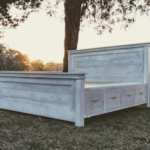 May include: A light gray wooden bed frame with a headboard, footboard, and side rails. The bed has three drawers built into the side. The bed is made of wood and has a distressed finish. The bed is outdoors on grass.