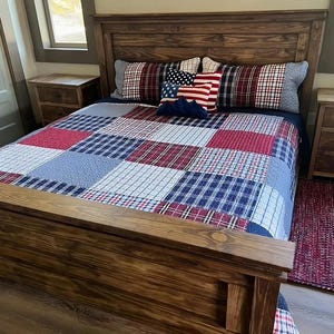 May include: A wooden bed frame with a patchwork quilt in red, white, and blue. The bed is made up with pillows featuring American flag and plaid patterns. A matching rug is on the floor.
