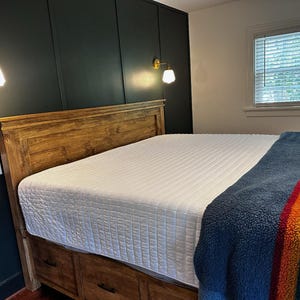 May include: A wooden bed frame with a white quilted mattress and a blue, striped blanket. The headboard is made of wood. Two gold sconces with white shades are mounted on the dark green wall.
