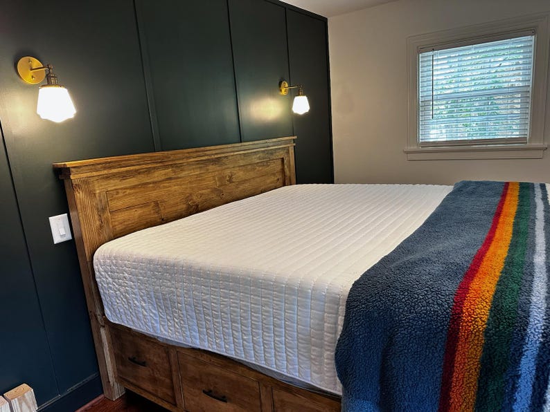 May include: A wooden bed frame with a white quilted mattress and a blue, striped blanket. The headboard is made of wood. Two gold sconces with white shades are mounted on the dark green wall.