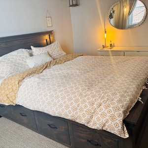 May include: A dark wood bed frame with built-in drawers, topped with a patterned duvet cover and pillows. The bedding features a white and gold floral design. A round mirror and dresser are visible in the background.