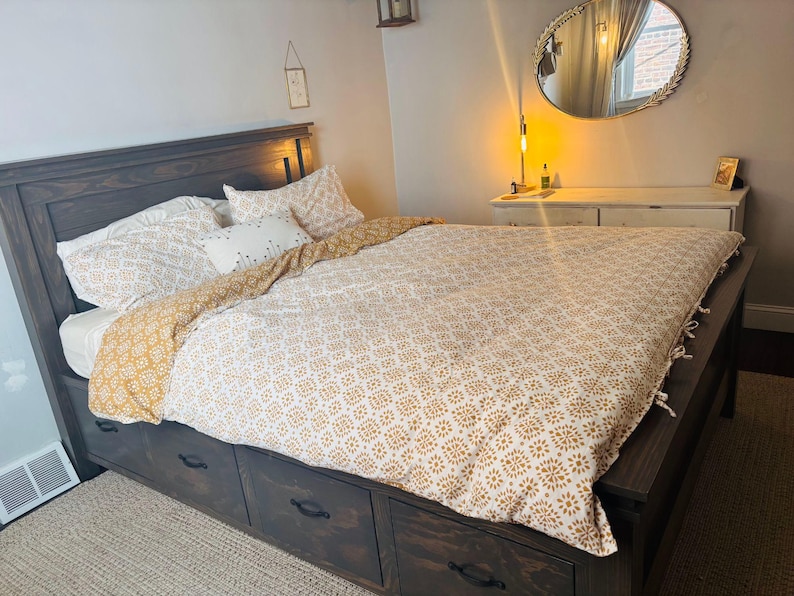 May include: A dark wood bed frame with built-in drawers, topped with a patterned duvet cover and pillows. The bedding features a white and gold floral design. A round mirror and dresser are visible in the background.