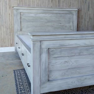 May include: A light gray wooden bed frame with a headboard and footboard. The bed frame has a distressed finish, with visible wood grain. The headboard is rectangular and the footboard has drawers. The bed is placed on a patterned rug.