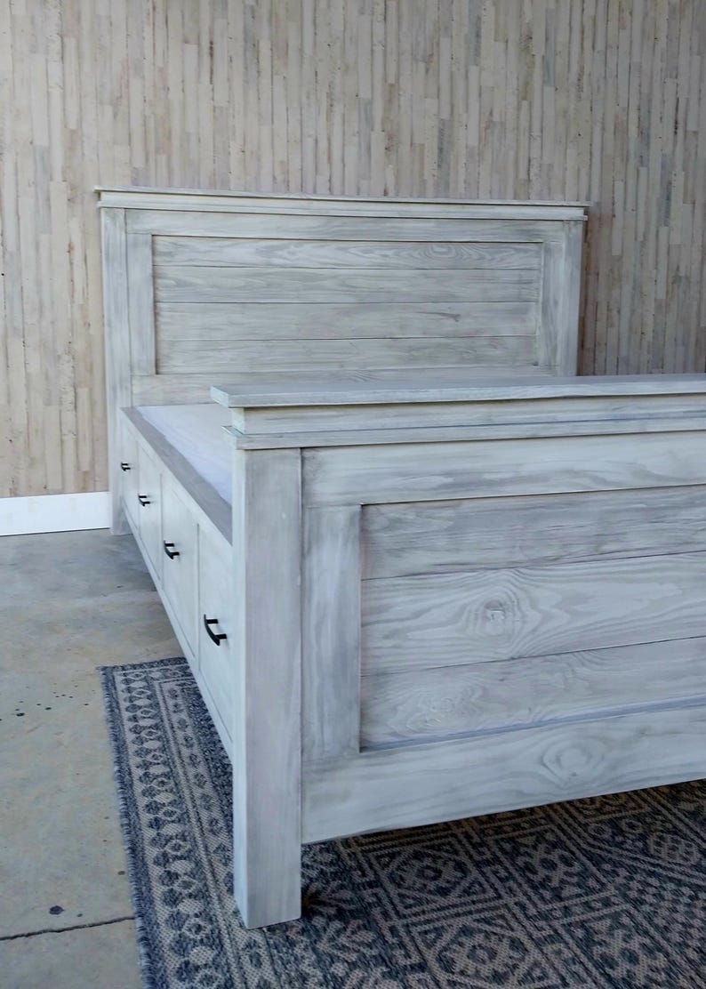May include: A light gray wooden bed frame with a headboard and footboard. The bed frame has a distressed finish, with visible wood grain. The headboard is rectangular and the footboard has drawers. The bed is placed on a patterned rug.