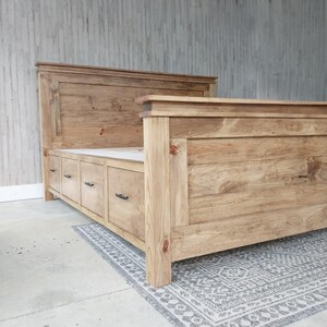 May include: A wooden bed frame with a headboard and footboard. The bed frame is made of light-colored wood and features a series of drawers built into the base. The headboard is rectangular and the footboard is lower. The bed is placed on a patterned rug.