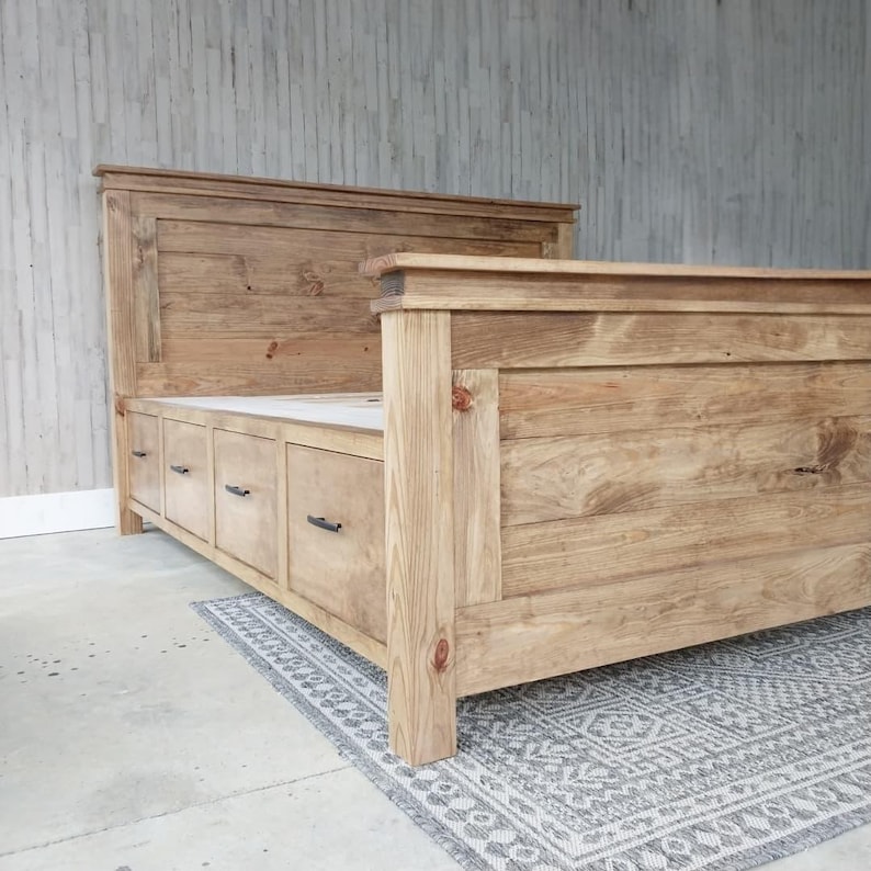May include: A wooden bed frame with a headboard and footboard. The bed frame is made of light-colored wood and features a series of drawers built into the base. The headboard is rectangular and the footboard is lower. The bed is placed on a patterned rug.