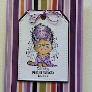 May include: A greeting card featuring a white tag with a stitched border, a purple and white twine bow, and a cartoon cat dressed as a bride with green eyes and a veil. The text reads Still looking FRIGHTENINGLY fabulous. The background has vertical stripes.