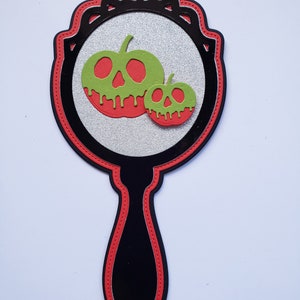 May include: A decorative hand mirror with a black handle and frame, accented with red trim. The mirror features a silver glitter background and two green and red apple-shaped designs, perfect for Halloween decor.