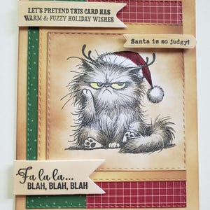 May include: A humorous Christmas card featuring an illustration of a grumpy cat wearing a Santa hat and antlers. Text includes "Let's pretend this card has warm & fuzzy holiday wishes", "Santa is so judgy!", and "Fa la la... BLAH, BLAH, BLAH". The card has a tan background with red and green accents.