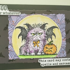 May include: A rectangular greeting card featuring a central circular illustration of a fluffy purple cat with black bat wings and a yellow bow, sitting next to an orange jack-o'-lantern. Text reads: BOO. Or whatever. This card may contain spells and sarcasm.