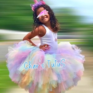 May include: A young person wearing a white tank top and a multi-colored tutu skirt. The tutu is pastel rainbow-colored. A unicorn horn headband is worn on the head. The text "Char's Tutus" is visible on the skirt.