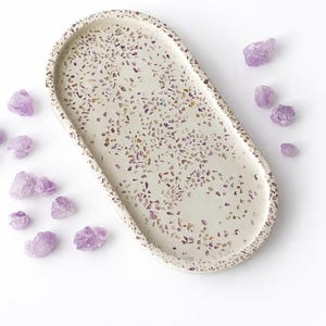 Puede incluir: Una bandeja ovalada de color blanquecino con un diseño moteado y cristales morados dispersos. La bandeja mide aproximadamente 30 cm de largo, perfecta para exhibir objetos pequeños o como pieza decorativa. Los cristales añaden un toque de belleza natural.