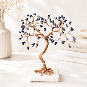 Op de afbeelding: Een decoratieve boomsculptuur met een stam en takken van gedraaid koperdraad, versierd met kleine, getrommelde blauwe lapis lazuli stenen. De boom is gemonteerd op een vierkante, witte stenen voet, wat zorgt voor een gebalanceerd en gestructureerd decoratieobject.