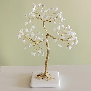 Könnte beinhalten: Eine goldene Drahtskulptur eines Baumes mit weißen Kristallsteinen als Blätter. Der Baum steht auf einem weißen Marmorsockel.