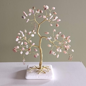 Puede incluir: Una escultura de árbol decorativa con ramas de alambre dorado trenzado adornadas con pequeñas piedras rosas pulidas. El árbol está montado sobre una base cuadrada blanca e incluye tres dijes de metal con las letras J, K y M.