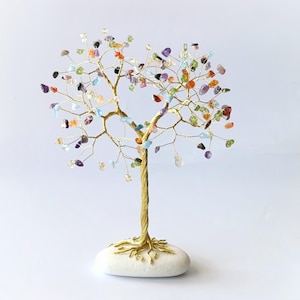 Puede incluir: Una escultura decorativa de árbol de alambre con un tronco y ramas de metal dorado trenzado, adornada con pequeñas piedras preciosas pulidas de varios colores en tonos morado, verde, azul, naranja y transparente, anclada sobre una base de piedra blanca.