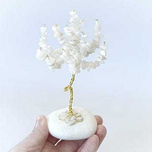 Op de afbeelding: Een decoratieve boomsculptuur gemaakt van witte stenen en goudkleurig draad. De boom is gemonteerd op een witte stenen basis. De sculptuur wordt in een hand gehouden tegen een witte achtergrond.