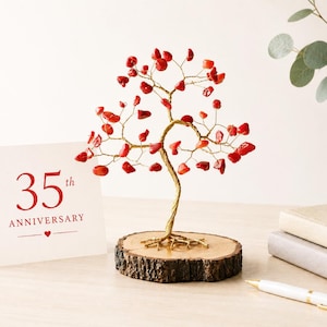 May include: A decorative wire tree with red gemstone leaves sits on a wooden base. A card with "35th Anniversary" in red lettering is next to the tree. A white pen and two notebooks are also on the table.