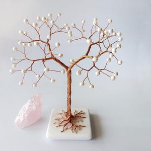 Puede incluir: Escultura de árbol hecha a mano con alambre de cobre y cuentas blancas perladas como hojas. El árbol está sobre una base cuadrada blanca con raíces de alambre de cobre. Un cristal de cuarzo rosa está junto al árbol.