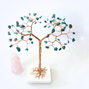May include: A handcrafted wire tree sculpture with turquoise-colored gemstone leaves. The copper-colored trunk and branches are affixed to a white square base. A pink rose quartz crystal sits beside the tree, creating a natural, decorative display.