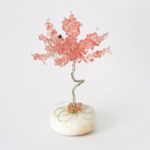 May include: A decorative tree sculpture with a silver wire trunk and branches. The tree's leaves are made of small, pinkish-red gemstones. The tree sits on a white stone base, with silver wire accents around the base.