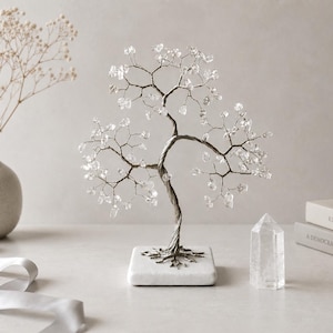 May include: A decorative wire tree sculpture featuring twisted silver-toned metal branches adorned with small, clear, translucent crystal chips. The tree is mounted on a square white marble base, creating a balanced and structured appearance.