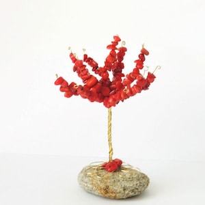 May include: A handcrafted decorative tree sculpture. The tree features a gold-colored wire trunk and branches adorned with vibrant red coral chips. The base is a natural gray stone, creating a unique and artistic display.