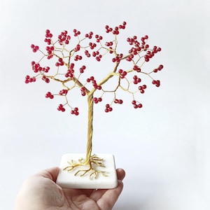 Op de afbeelding: Handgemaakte draadboomsculptuur met een goudkleurige stam en takken, versierd met rode kralen. De boom staat op een witte vierkante basis. De sculptuur wordt in een hand gehouden tegen een witte achtergrond.