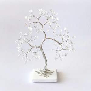 May include: A decorative tree sculpture with a silver-colored wire trunk and branches. The branches are adorned with clear, faceted crystal-like stones. The tree sits on a square, white base, creating a unique home decor item.