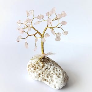 Op de afbeelding: Een decoratieve boomsculptuur met een goudkleurige draadstam en takken, versierd met roze edelsteenbladeren. De boom staat op een witte, poreuze stenen basis, met een klein lint en het nummer "2026".