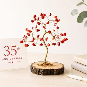 Può includere: Un albero decorativo in filo metallico con foglie di pietre preziose rosse su una base di legno. Una carta con "35° Anniversario" in lettere rosse è accanto all'albero. Una penna bianca e due quaderni sono anche sul tavolo.