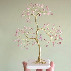 May include: A handcrafted wire tree sculpture with a gold-colored trunk and branches, adorned with pink gemstone 'leaves'. A pink ribbon charm hangs from a branch. The tree sits on a white square base.