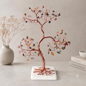 Puede incluir: Una escultura decorativa de árbol hecha de alambre trenzado color cobre, con numerosas pequeñas piedras preciosas multicolores unidas a las ramas. El árbol está montado sobre una base cuadrada de piedra blanca con detalles de raíces visibles.