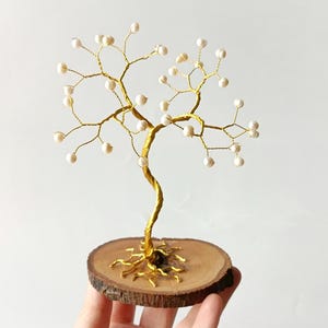 Könnte beinhalten: Handgefertigte Baumskulptur aus goldfarbenem Draht und kleinen, weißen Perlen. Der Baum steht auf einem runden Holzsockel mit natürlichem Rand. Ein einzigartiges, handwerkliches Objekt.