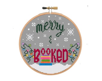 May include: A cross-stitch embroidery hoop with the words "merry & booked" in dark green and red. The design includes snowflakes, a stack of books, and string lights. The background is gray, and the hoop is made of wood.