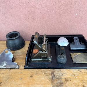 Puede incluir: Un surtido de suministros de oficina antiguos, que incluyen un tintero negro, un clip de metal, una perforadora, una grapadora y un portapapeles, todos dispuestos sobre una superficie de madera. La grapadora tiene una tapa plateada con la palabra "EXCEL" impresa.