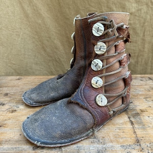 Puede incluir: Un par de botas de cuero marrón desgastado, con cierre de botones y cordones. Las botas tienen una punta redondeada y están cosidas. El cuero muestra signos de desgaste y envejecimiento, con una estética rústica.