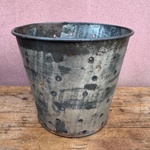 May include: A weathered, galvanized metal bucket with numerous small holes. The bucket has a slightly tapered shape and a dark rim. The metal shows signs of age and wear, with areas of discoloration and rust. The bucket sits on a wooden surface.