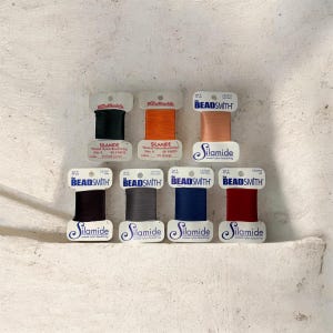 May include: A collection of eight spools of Beadsmith Silamide waxed nylon beading thread in various colors, including black, orange, and burgundy. Each spool is labeled with the thread's color and size, and is approximately 40 yards in length.
