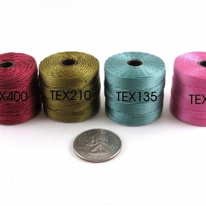 Op de afbeelding: Vier spoelen garen in verschillende kleuren: bordeauxrood, olijfgroen, teal en roze. Elke spoel is gelabeld met de tekst "TEX" gevolgd door een nummer. Een Amerikaanse kwart is getoond voor schaal.