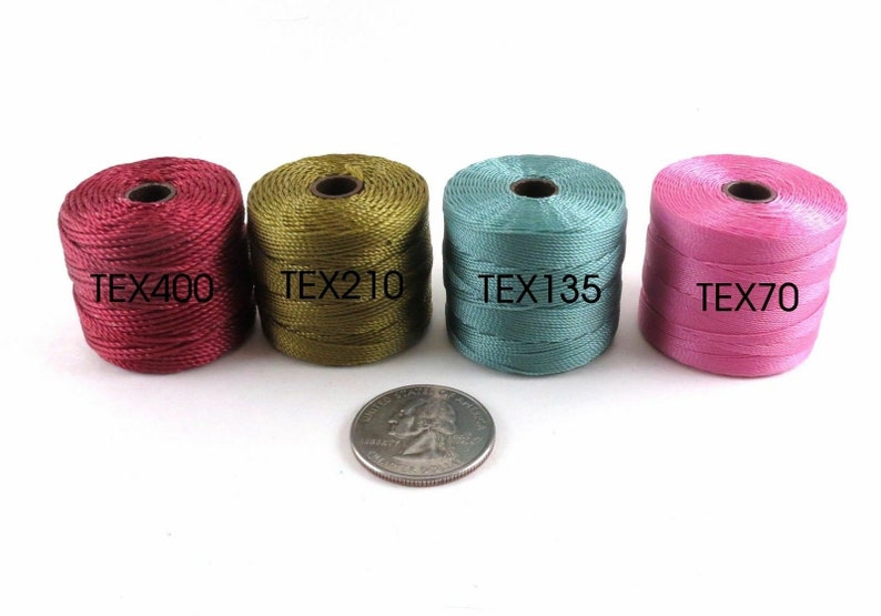 Op de afbeelding: Vier spoelen garen in verschillende kleuren: bordeauxrood, olijfgroen, teal en roze. Elke spoel is gelabeld met de tekst "TEX" gevolgd door een nummer. Een Amerikaanse kwart is getoond voor schaal.