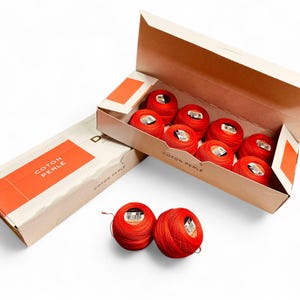 Op de afbeelding: Een open en een gesloten doos met rood katoenen parelgaren. De open doos bevat acht bollen garen. Een gesloten doos staat ernaast. Twee extra bollen rood garen liggen voor de dozen. De dozen hebben oranje accenten en de tekst "COTON PERLE".