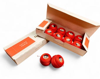 Puede incluir: Cajas de hilo de algodón perlado rojo, una abierta y otra cerrada. La caja abierta contiene ocho ovillos de hilo. Dos ovillos de hilo rojo adicionales están delante de las cajas. Las cajas tienen detalles en naranja y el texto "COTON PERLE".
