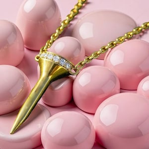 May include: A gold-colored pendant necklace with a diamond-studded top and a long, pointed design. The necklace is set against a backdrop of pink, glossy, egg-shaped objects. The chain is also gold-colored.
