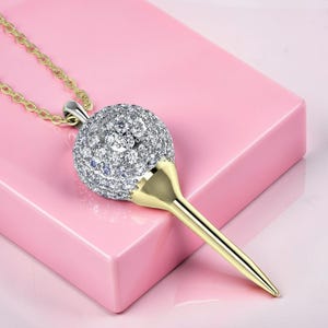 May include: A gold-toned pendant necklace featuring a golf ball design. The golf ball is encrusted with small, sparkling stones. The pendant has a gold-colored tee and chain, set against a pink background.