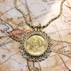 May include: A gold-toned necklace featuring a pendant with an 1814 coin set within a decorative filigree frame. The necklace rests on a vintage map, showcasing a detailed design and a classic aesthetic.