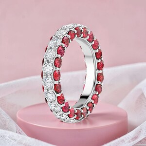 May include: A silver ring with two rows of gemstones. One row features bright red gemstones, and the other row features clear, white gemstones. The ring is set against a soft pink background.