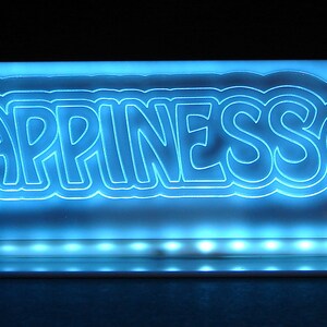 May include: A blue neon sign displaying the word "HAPPINESS" in a playful font, accompanied by a smiling face. The sign is illuminated with a bright blue glow, and has a rectangular base.