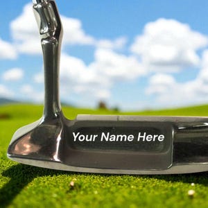 May include: A shiny silver golf putter with the text "Your Name Here" on the club head. The putter is set against a backdrop of green grass, a golf course, and a blue sky with fluffy white clouds.